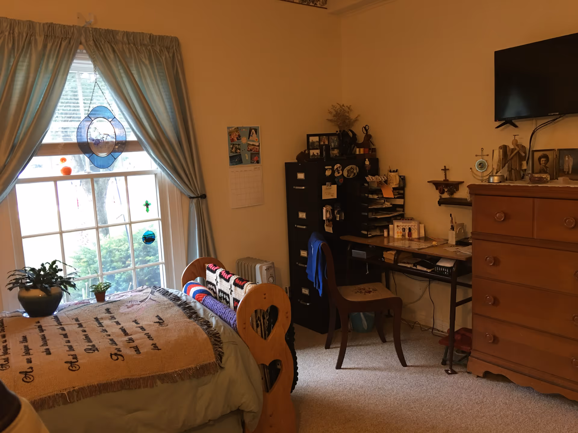 A cozy bedroom with a single bed covered with a blanket featuring text. The bed is positioned near a large window with light blue curtains and decorative hanging ornaments. There are two potted plants on the windowsill. To the right of the bed is a wooden dresser with a flat-screen TV mounted above it. Next to the dresser is a desk with a chair, a filing cabinet, and various personal items and decorations on the desk and walls.