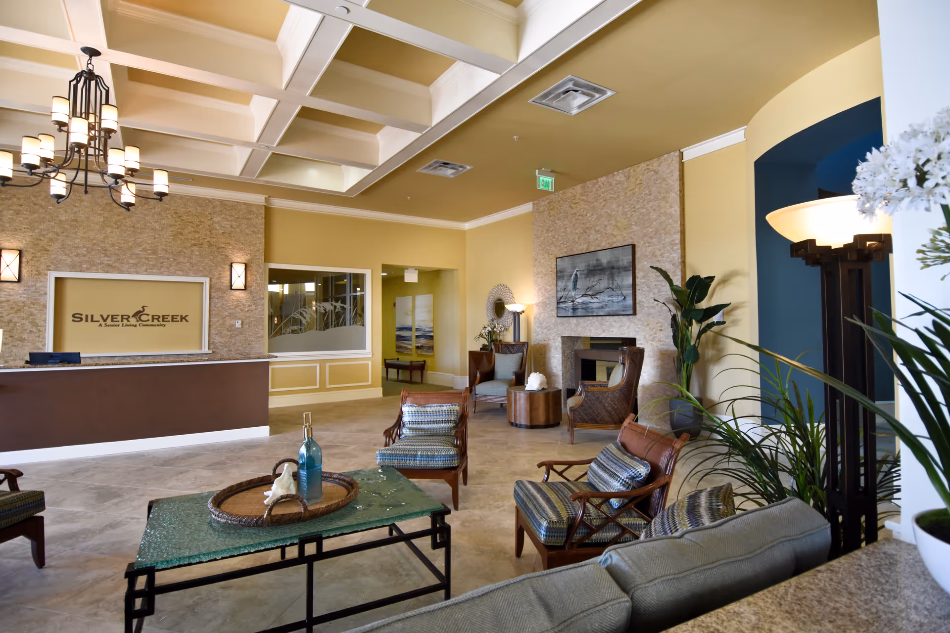 A spacious and well-lit senior living community lobby with a reception desk on the left featuring the Silver Creek logo. The room has a high coffered ceiling with a chandelier, comfortable seating including armchairs and a sofa around a glass coffee table, decorative plants, and a fireplace with artwork above it.