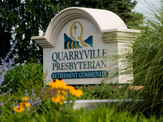 A white stone sign with an arched top displaying the logo and name Quarryville Presbyterian Retirement Community, surrounded by green plants and yellow flowers.