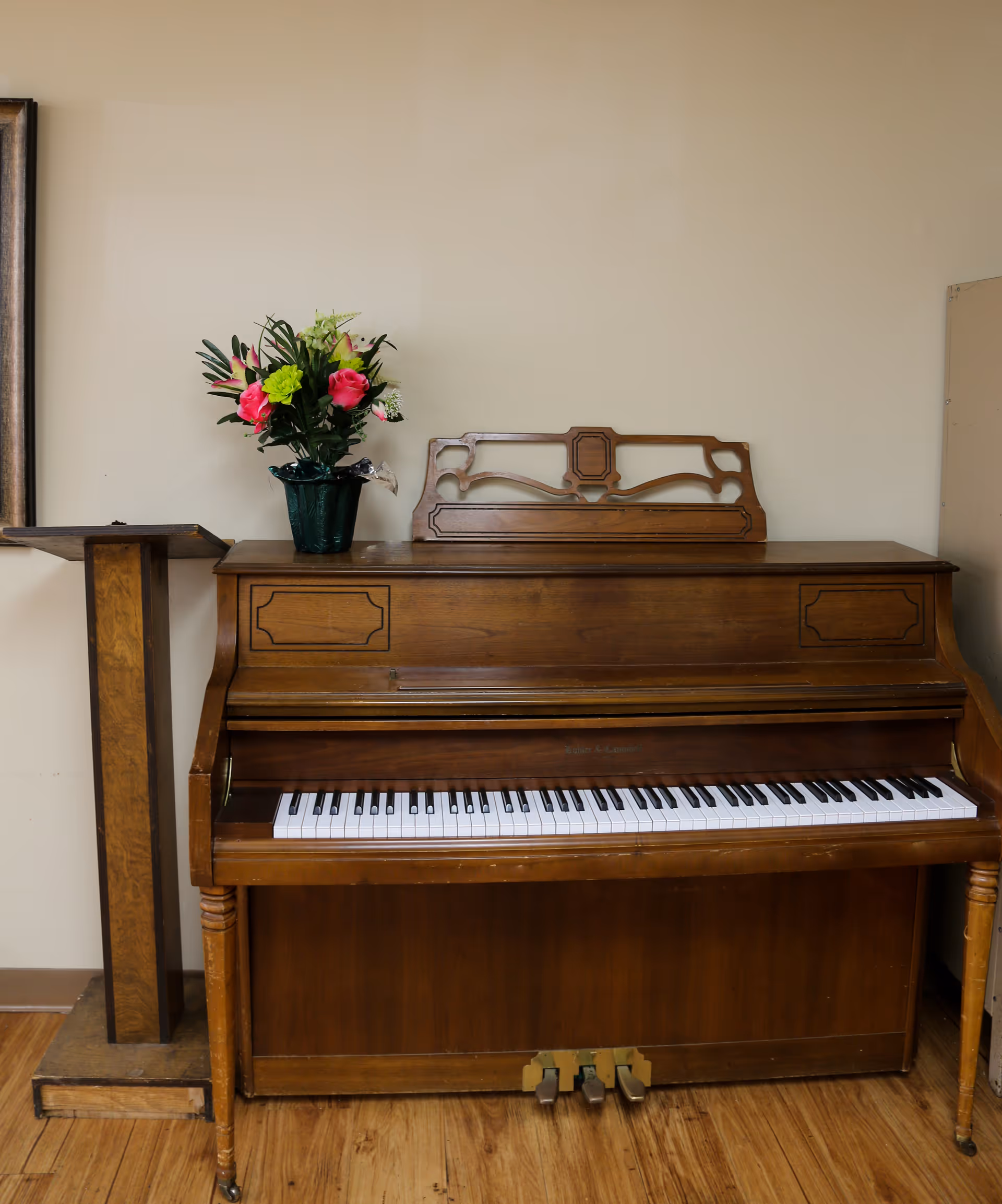 An upright wooden piano with a decorative music stand on top, placed against a beige wall. A green vase with colorful artificial flowers sits on the piano. To the left of the piano is a wooden lectern. The floor is wooden.