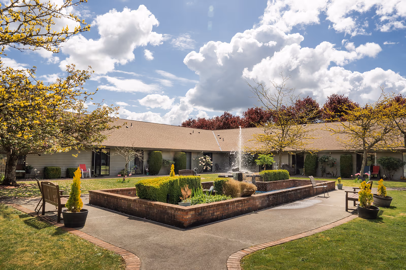 Outdoor courtyard area at Village Concepts of Marysville - Grandview Village featuring a central brick planter with a water fountain, surrounded by benches and potted plants. Trees with yellow and red foliage frame the courtyard under a partly cloudy blue sky.