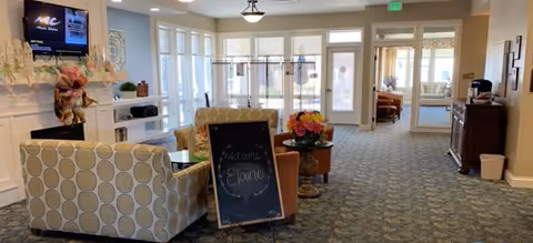 Well-lit common area with upholstered seating, a chalkboard welcome sign, a TV on the wall, and glass doors at the far end.