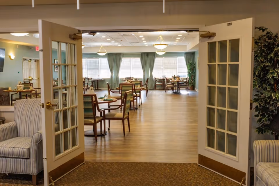 View through open double doors into a spacious dining room with multiple tables and chairs arranged neatly. The room has large windows with green curtains allowing natural light to fill the space. There are two striped armchairs positioned near the doors and a large potted plant on the right side.
