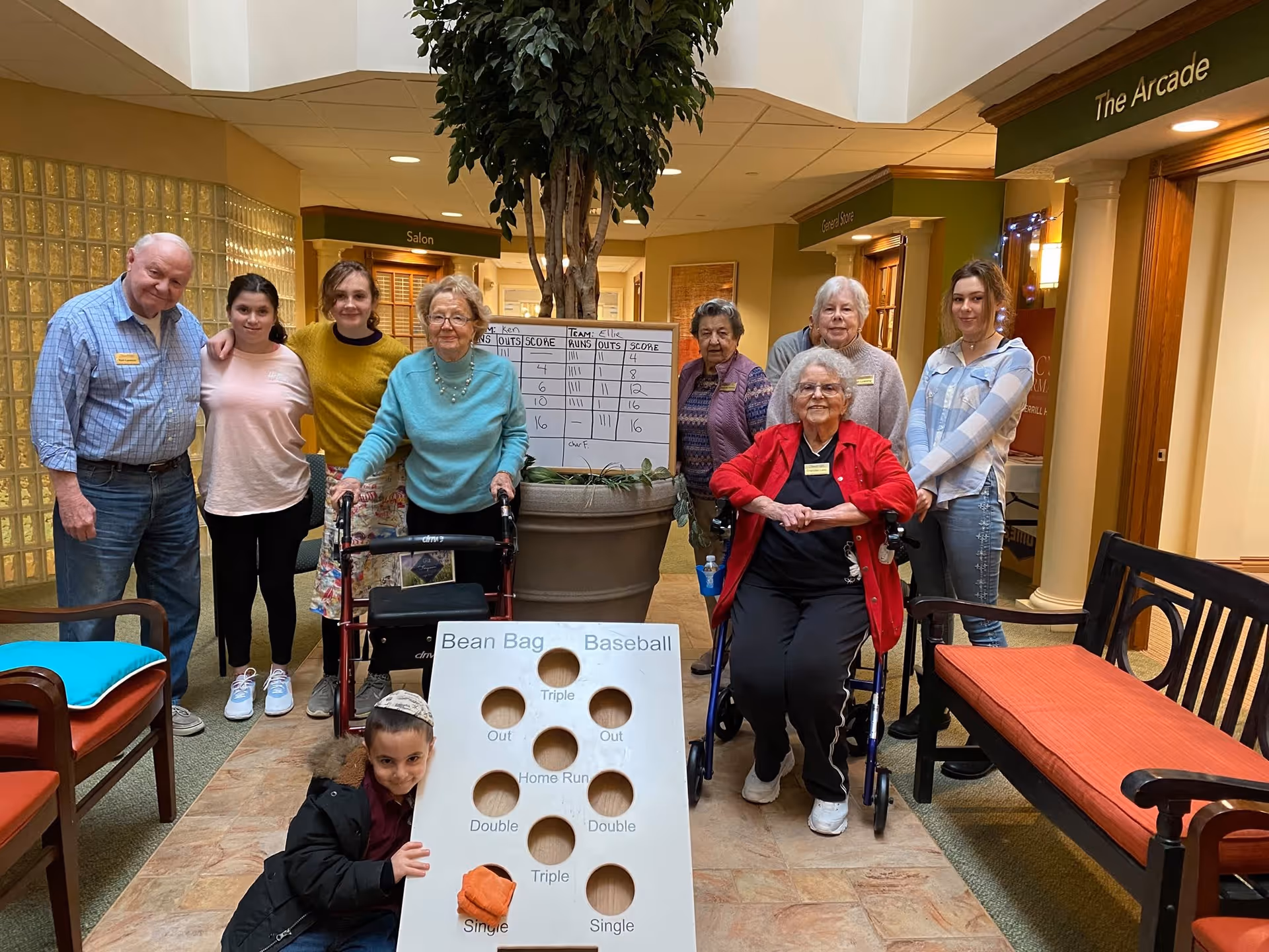 A group of elderly residents and younger individuals posing together in a well-lit indoor common area of a retirement facility. Two elderly women are seated in walkers, while others stand behind them. In front of the group is a bean bag baseball game board with holes labeled for different baseball plays. The area has benches with colorful cushions and signs indicating nearby rooms such as 'Salon' and 'The Arcade'.