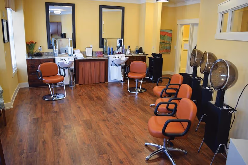 Interior view of a salon area with wooden flooring, yellow walls, and multiple orange salon chairs. There are two mirrors above counters with sinks, hair care products, and towels. On the right side, there are three hair dryers with chairs underneath them. A window and a painting are visible on the walls.