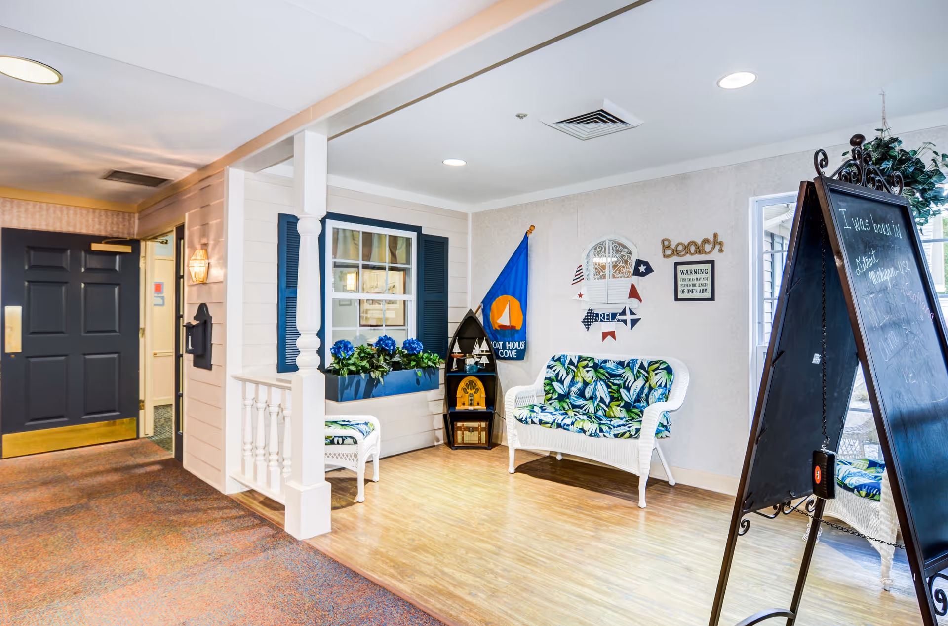 A cozy indoor seating area with a white wicker loveseat and chair featuring blue and green leaf-patterned cushions. The wall behind has nautical-themed decorations including a blue flag with a sailboat, a sign that says 'Beach', and a warning sign about fish tales. There is a blackboard on a stand with handwritten text, a window with blue shutters and a flower box with blue flowers, and a dark door with a brass handle in the background.