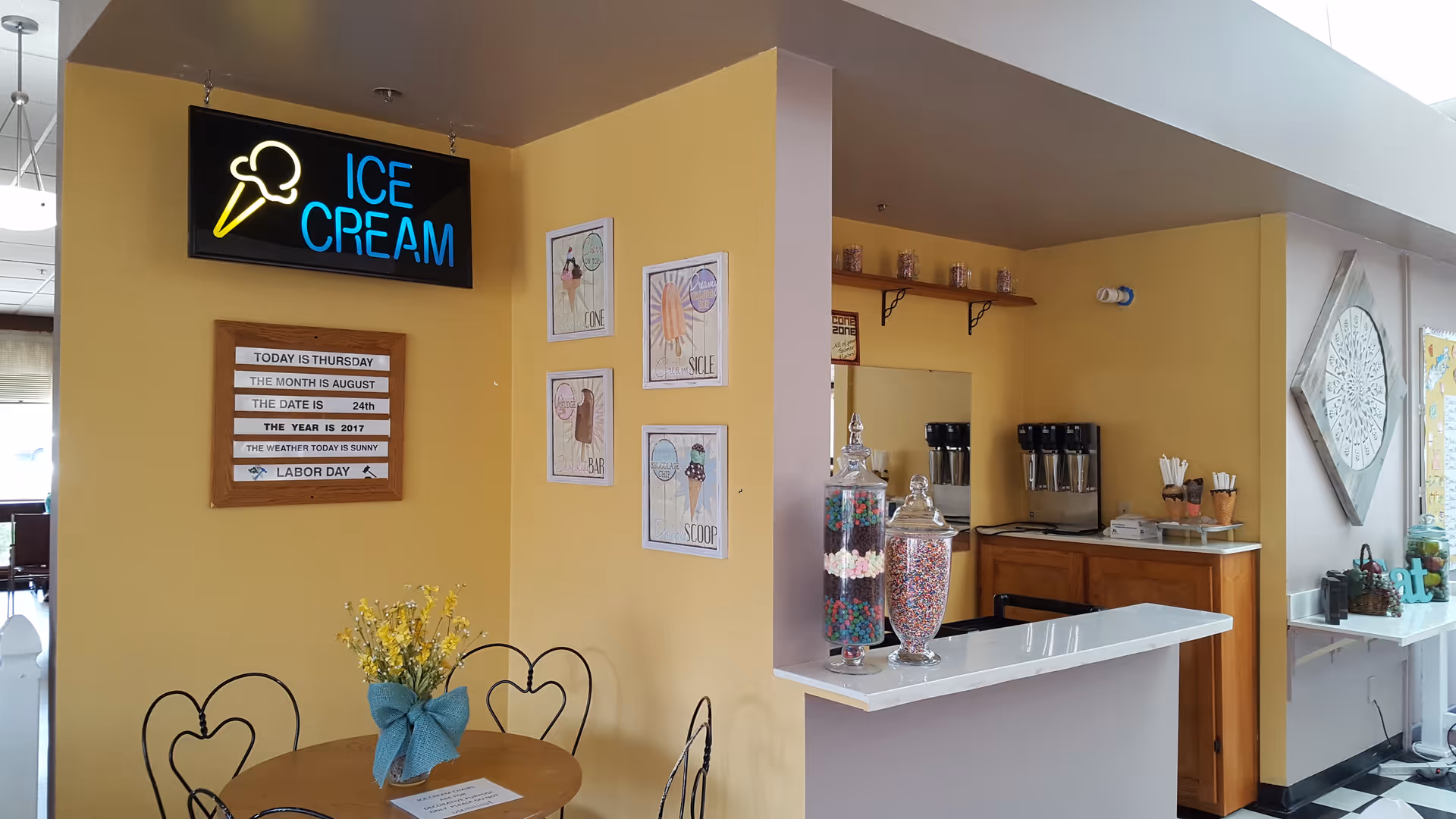 Interior view of an ice cream corner in a senior living facility with a round table and chairs, a vase with yellow flowers tied with a blue ribbon on the table, a neon sign reading 'ICE CREAM' with an ice cream cone icon above it, a wooden board displaying the date and weather, framed ice cream-themed artwork on the yellow walls, a counter with jars of colorful candies, and a coffee station with dispensers and cones on a wooden cabinet.