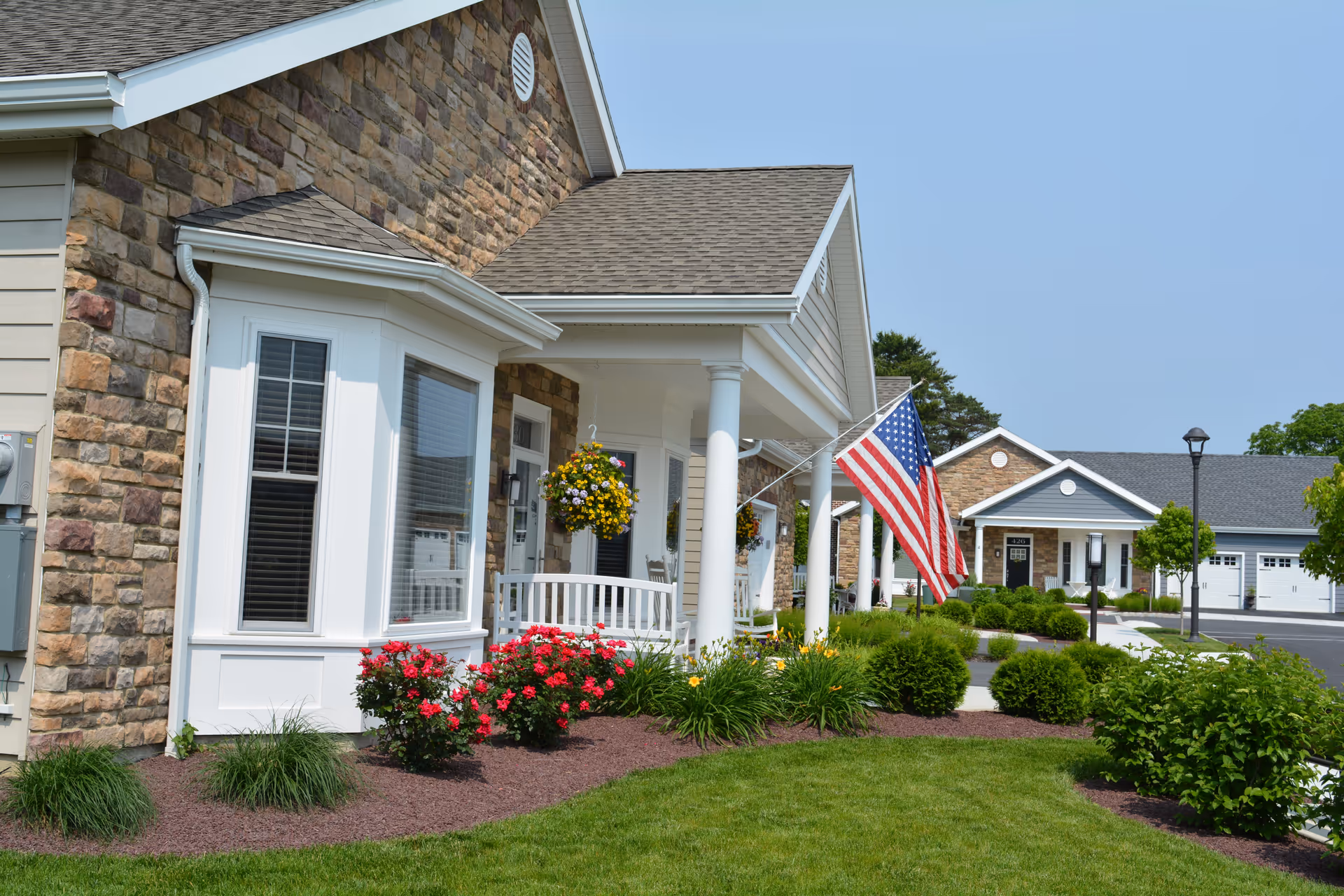 Exterior view of a senior living facility with stone and siding walls, white columns supporting a porch roof, hanging flower baskets, an American flag, well-maintained landscaping with bushes and flowers, and a paved driveway with garages in the background under a clear blue sky.