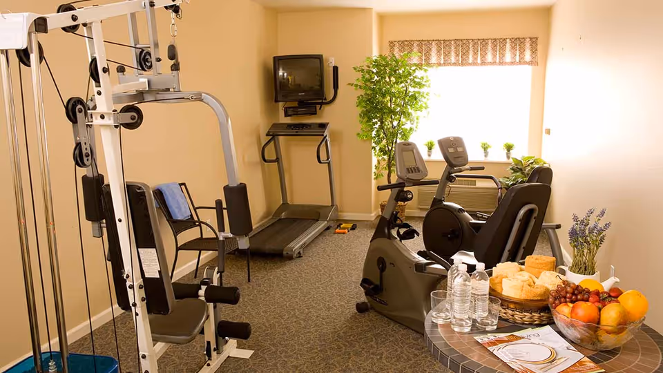 A small fitness room with exercise equipment including a multi-gym machine, a treadmill, and two stationary bikes. There is a TV mounted on the wall above the treadmill. A large window with a valance lets in natural light, and there are several potted plants on the windowsill and floor. In the foreground, a round table holds a bowl of fresh fruit, a basket of towels, water bottles, glasses, and some brochures. The room has beige walls and carpeted flooring.