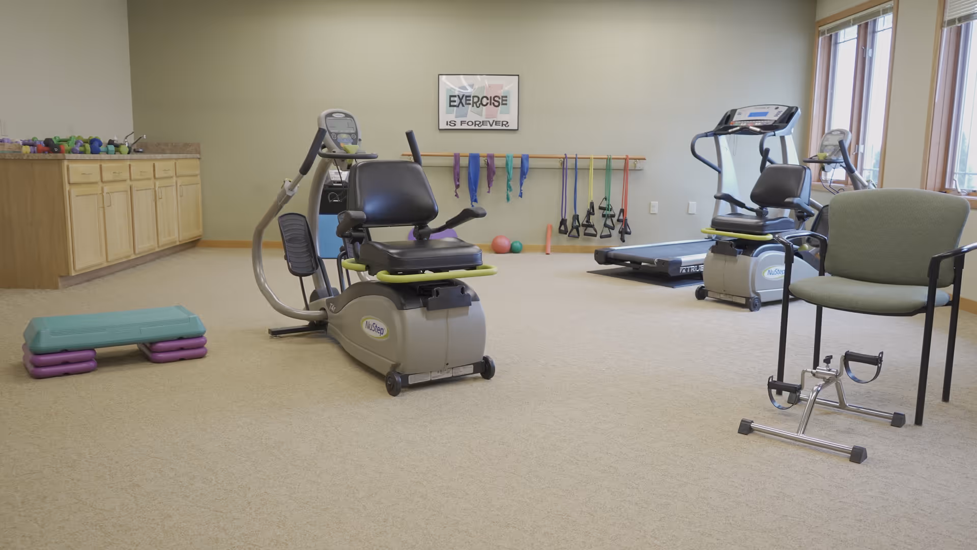 A well-lit exercise room with various fitness equipment including a NuStep recumbent cross trainer, a treadmill, resistance bands hanging on the wall, exercise balls, a step platform, and a pedal exerciser next to a green chair. There is a countertop with cabinets along one wall and a framed sign on the wall that reads 'EXERCISE IS FOREVER'.