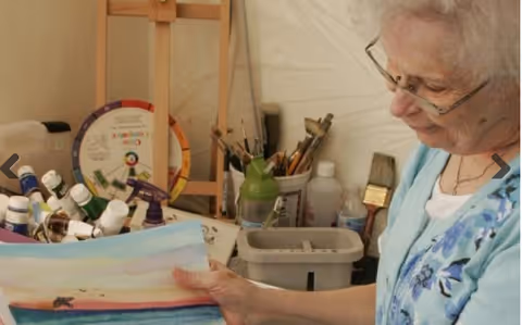 An elderly woman with glasses is sitting and holding a painting she created. She is in a room with various art supplies including paint bottles, brushes, and a color wheel on a stand behind her.