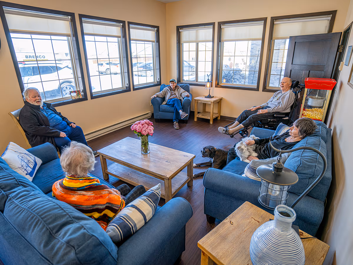 Several residents seated on blue sofas and chairs around a wooden coffee table in a bright common living room.
