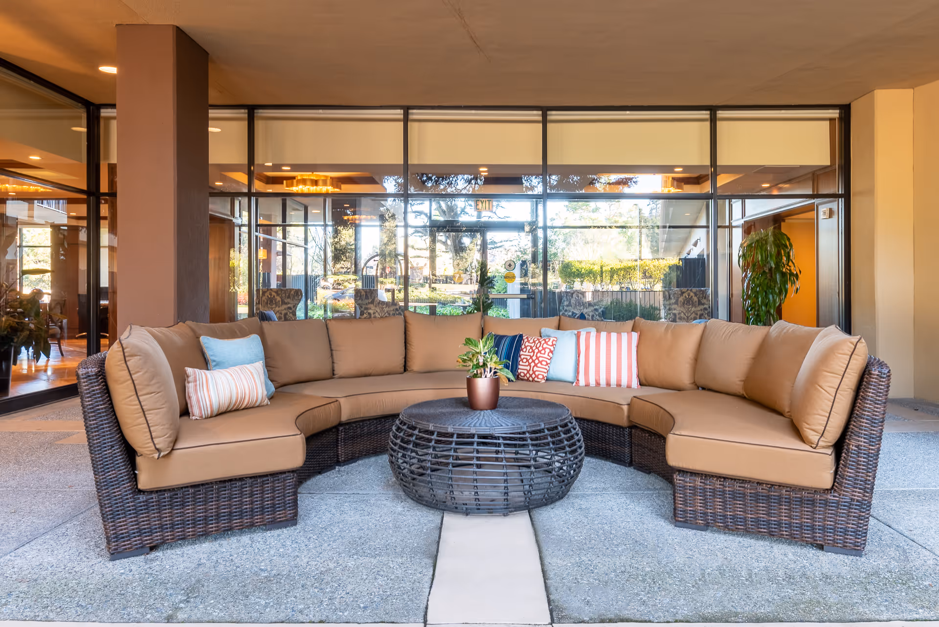 Curved wicker seating with tan cushions and colorful pillows around a round wicker coffee table in a covered entrance patio in front of glass doors.