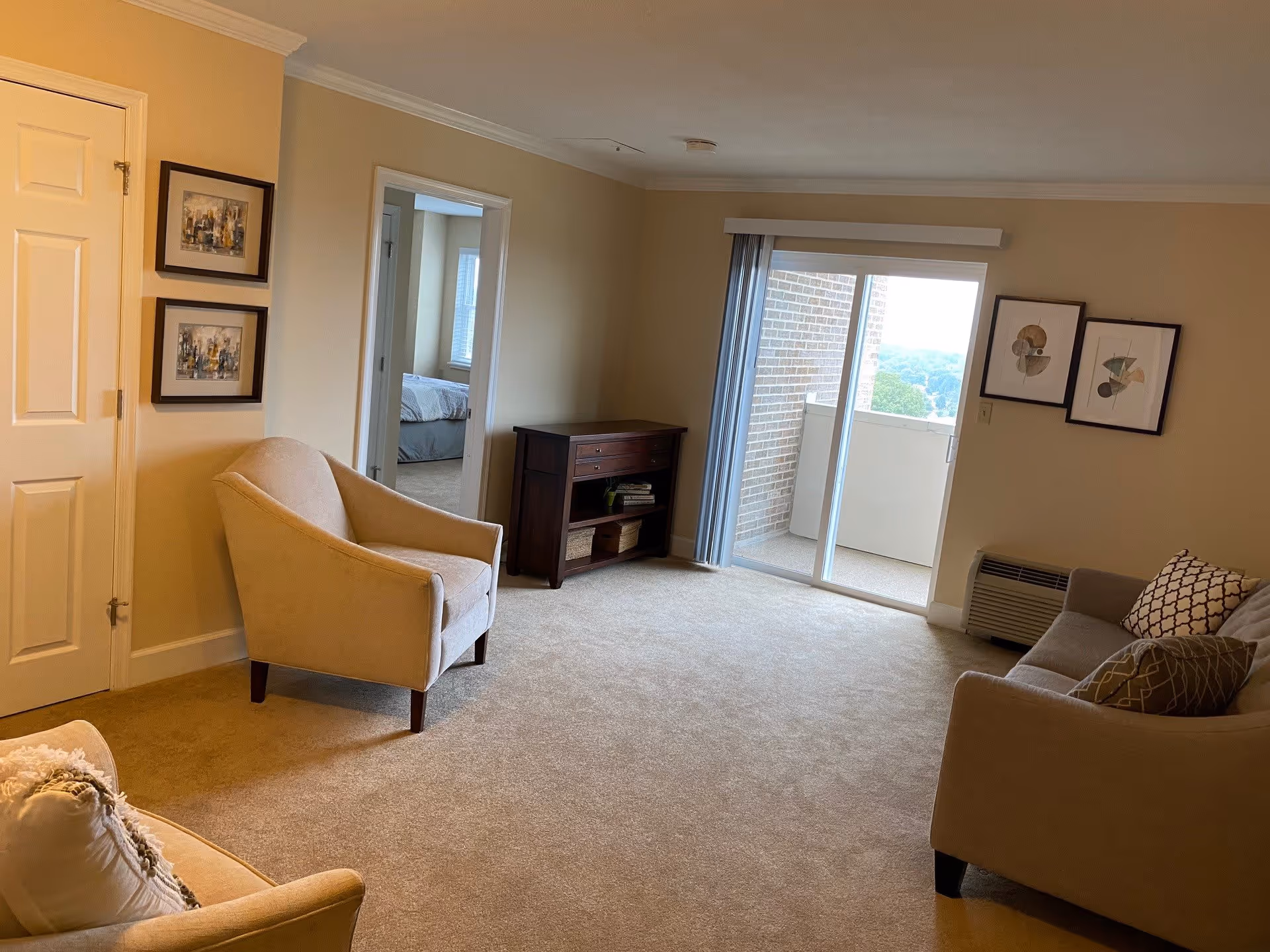 A living room with beige carpet and walls, featuring a beige armchair and a gray sofa with patterned pillows. There is a small dark wooden cabinet against the wall, two framed abstract artworks hanging above a heating unit, and a sliding glass door leading to a balcony. A doorway leads to a bedroom with a bed visible in the background.