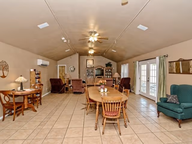 A spacious senior living common area with a long wooden dining table and chairs in the center. The room features tiled floors, beige walls, ceiling fans with lights, and multiple seating options including armchairs and a green upholstered chair. There are French doors with curtains on the right side, a wooden cabinet with a TV and decor at the far end, and a small round table with chairs and a lamp on the left side.