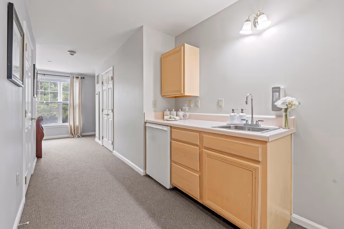 A hallway with a small kitchenette on the right side featuring a sink, countertop, wooden cabinets, and a mini refrigerator. At the end of the hallway, there is a window with beige curtains allowing natural light to enter. The walls are painted light gray and the floor is carpeted in a neutral tone.