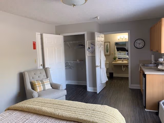 Interior view of a senior living facility room at Summit Place showing a bed with a yellow blanket in the foreground, a gray armchair with a plaid pillow, an open closet with hangers, a bathroom with a sink and mirror visible through an open door, a wall clock, and a kitchenette area with wooden cabinets and a countertop.