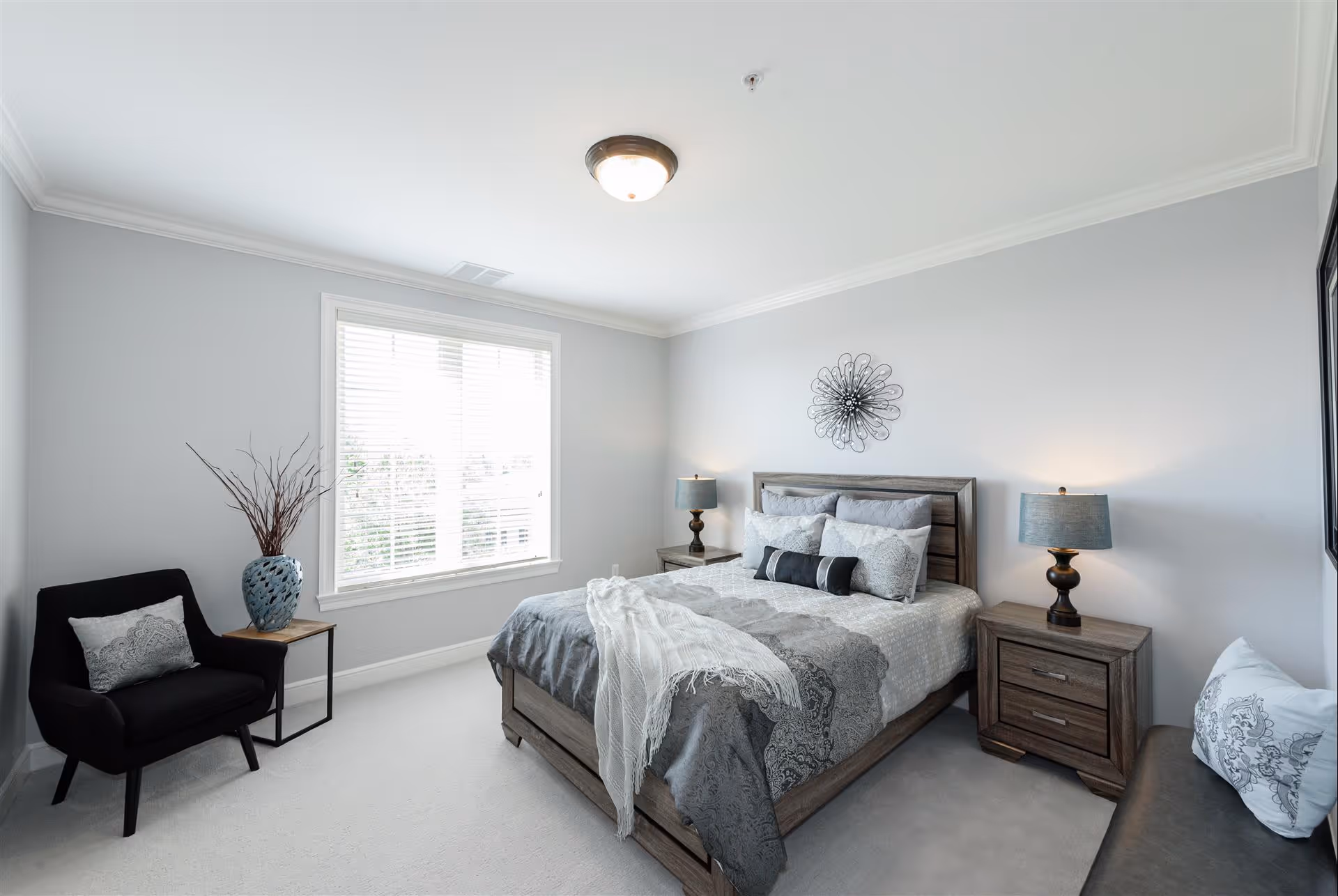 A bright and neatly arranged bedroom with a large window letting in natural light. The room features a wooden bed with gray and white bedding, two matching nightstands with lamps, a black armchair with a decorative pillow, a small side table with a vase of branches, and a cushioned bench with a pillow. The walls are painted light gray and there is a decorative metal wall art above the bed.