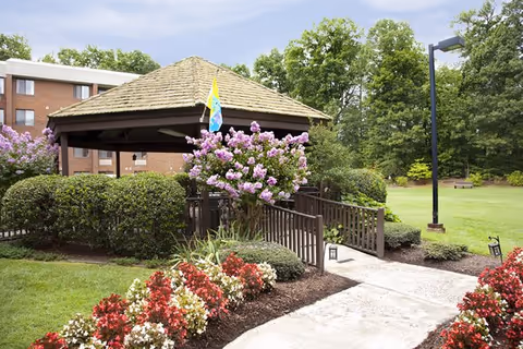 A landscaped outdoor area featuring a wooden gazebo with a shingled roof surrounded by flowering bushes and colorful flower beds. A paved walkway leads to the gazebo, and there are trees and a grassy lawn in the background. A multi-story brick building is visible behind the gazebo.