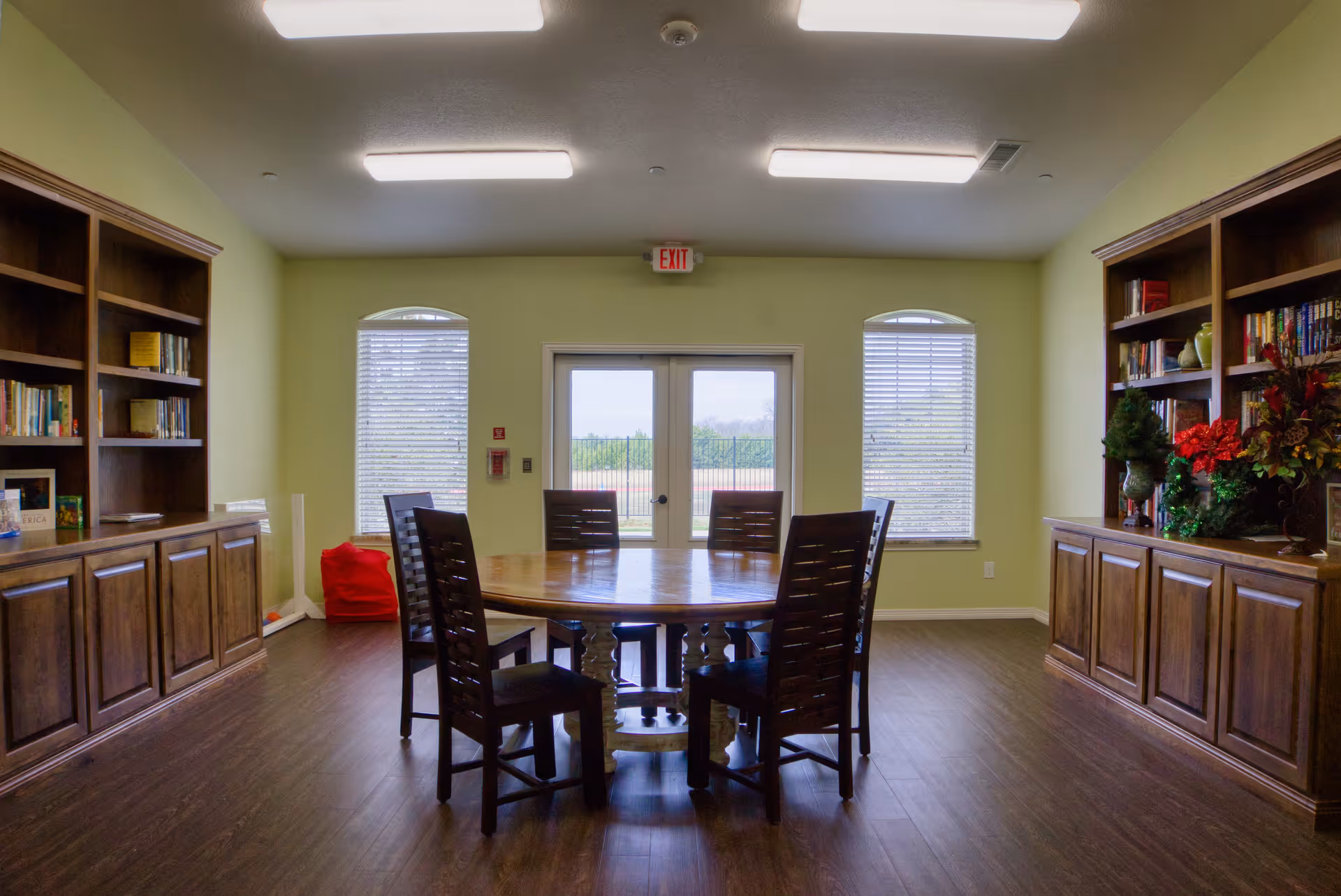A well-lit room with a round wooden table surrounded by six chairs in the center. The room has wooden bookshelves filled with books and decorative plants on both sides. Two windows with blinds and a glass door leading outside are visible at the far end. The floor is wooden, and the walls are painted light green.