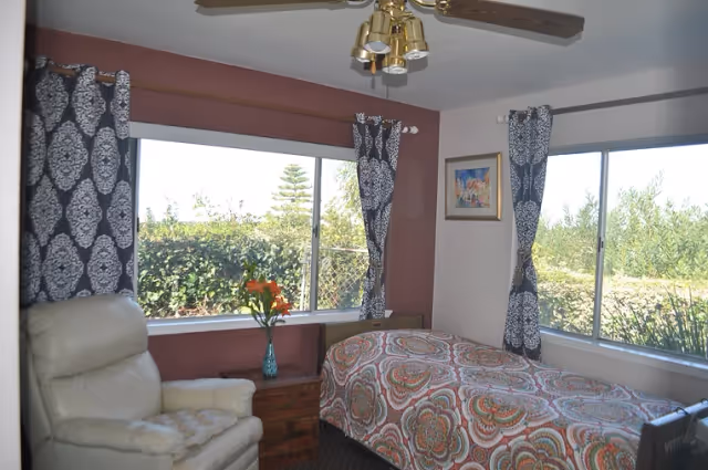 A cozy bedroom with a single bed covered in a colorful patterned bedspread. There are two large windows with black and white patterned curtains, letting in natural light and showing greenery outside. Next to the bed is a wooden nightstand with a vase of orange flowers. A cream-colored armchair is positioned near the window, and a ceiling fan with brass light fixtures is visible above. A framed picture hangs on the wall.
