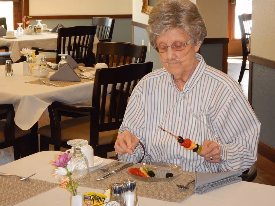 An elderly woman wearing a nasal cannula sits at a dining table assembling a fruit skewer in a dining room.