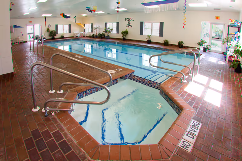 Indoor pool and adjacent spa in a tiled recreation room with handrails, plants, and a 'Pool & Spa' sign on the wall.