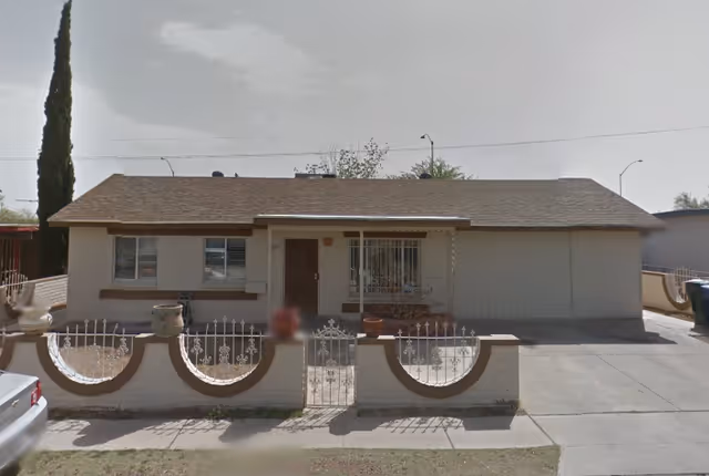 Single-story residential building with a brown shingle roof and beige exterior walls. The front yard is enclosed by a low white wall with decorative iron fencing and a small gate. There are two windows on the left side and a door in the center with a small covered porch area. A driveway is visible on the right side of the house, and a tall tree is on the left side of the yard.