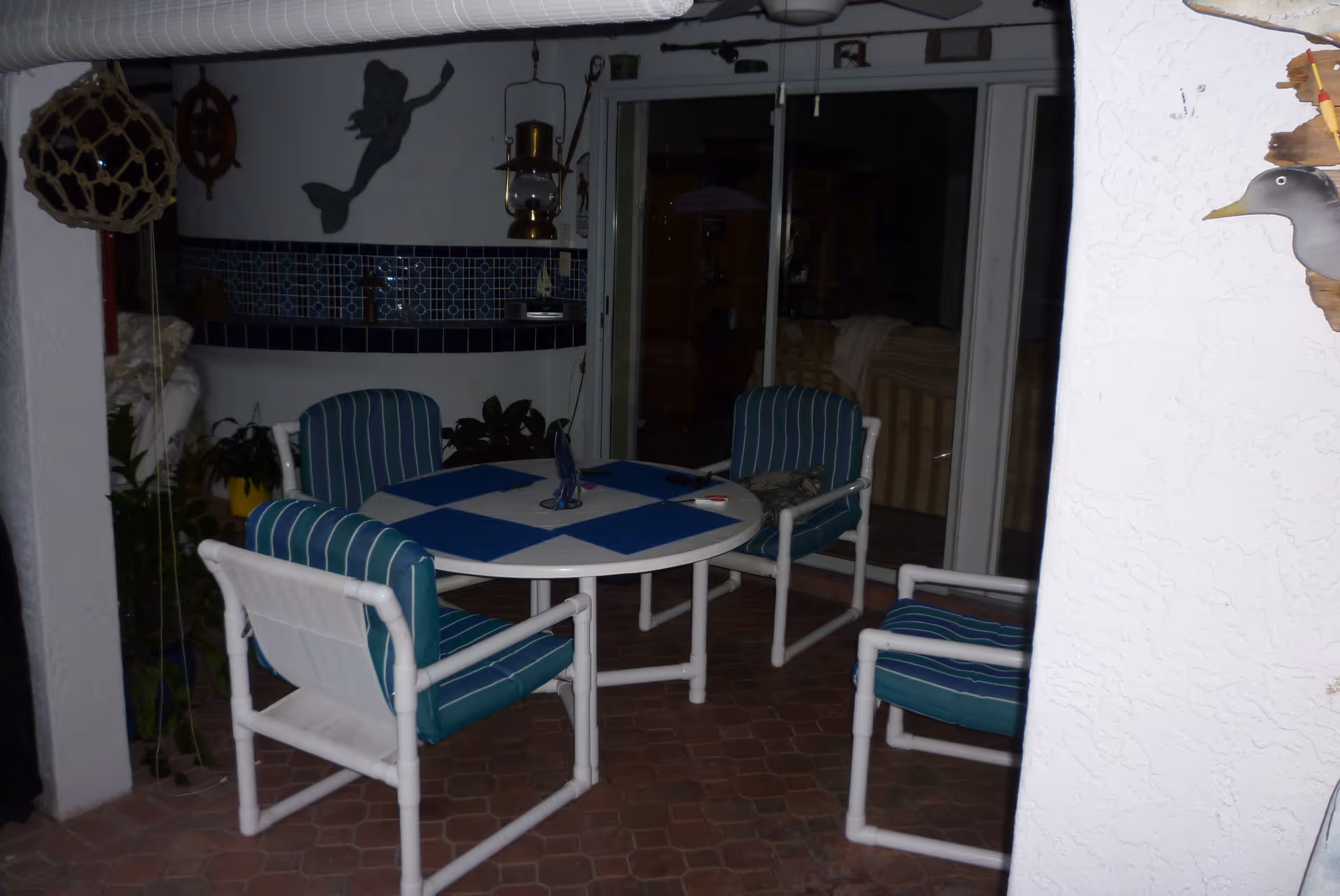 Round patio table surrounded by four blue-striped cushioned chairs on a tiled covered porch with sliding glass doors and nautical decor.
