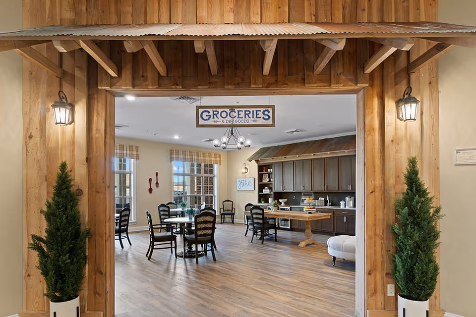 Interior view of a rustic-style dining and common area in an assisted living facility. The room features wooden floors, a wooden beam entrance with two potted plants on either side, and a hanging sign that reads 'GROCERIES & DRY GOODS'. Inside, there are several tables and chairs, a large wooden table with food items, cabinetry along the back wall, and windows with plaid valances letting in natural light.