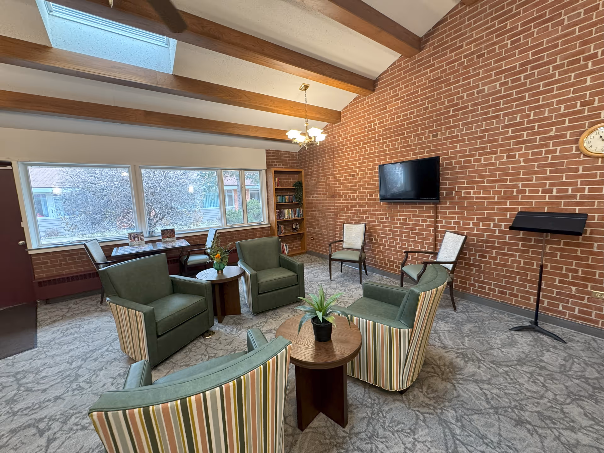 Cozy senior living lounge with green armchairs arranged around small tables, a wall-mounted TV on exposed brick, large windows and a skylight.
