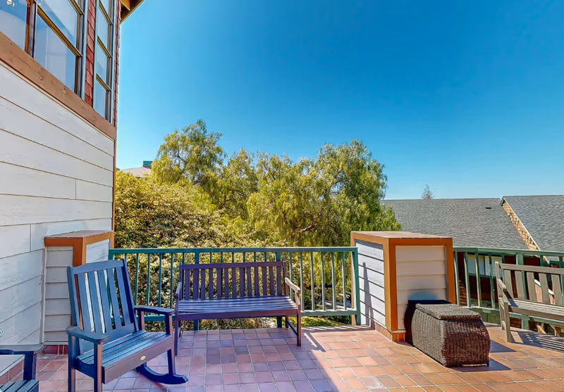 Outdoor balcony with wooden chairs and a bench overlooking trees under a clear blue sky.