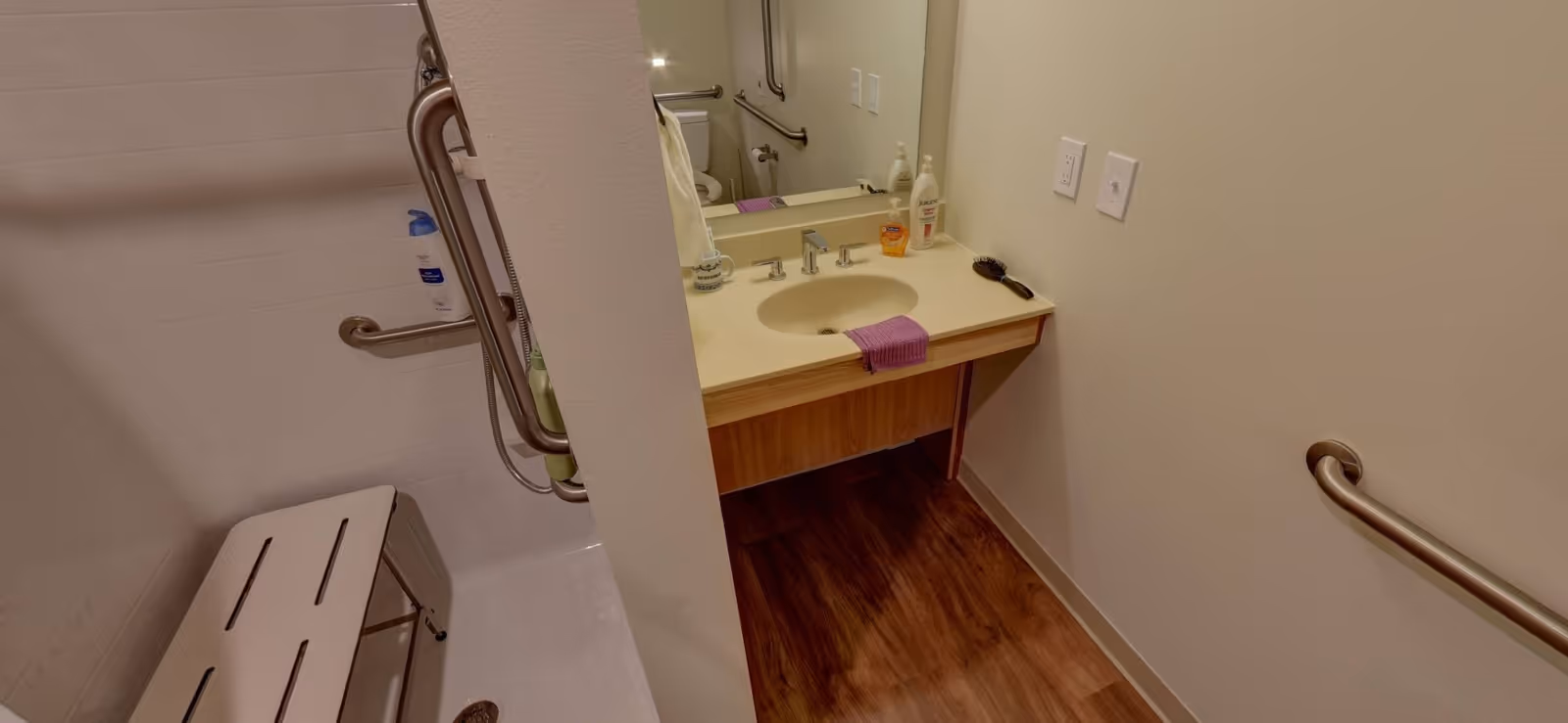 A bathroom with a shower area featuring a white shower bench and stainless steel grab bars. The vanity has a sink with a purple hand towel, soap dispenser, lotion, and a hairbrush on the countertop. The floor is wood-style, and there is a large mirror above the sink.