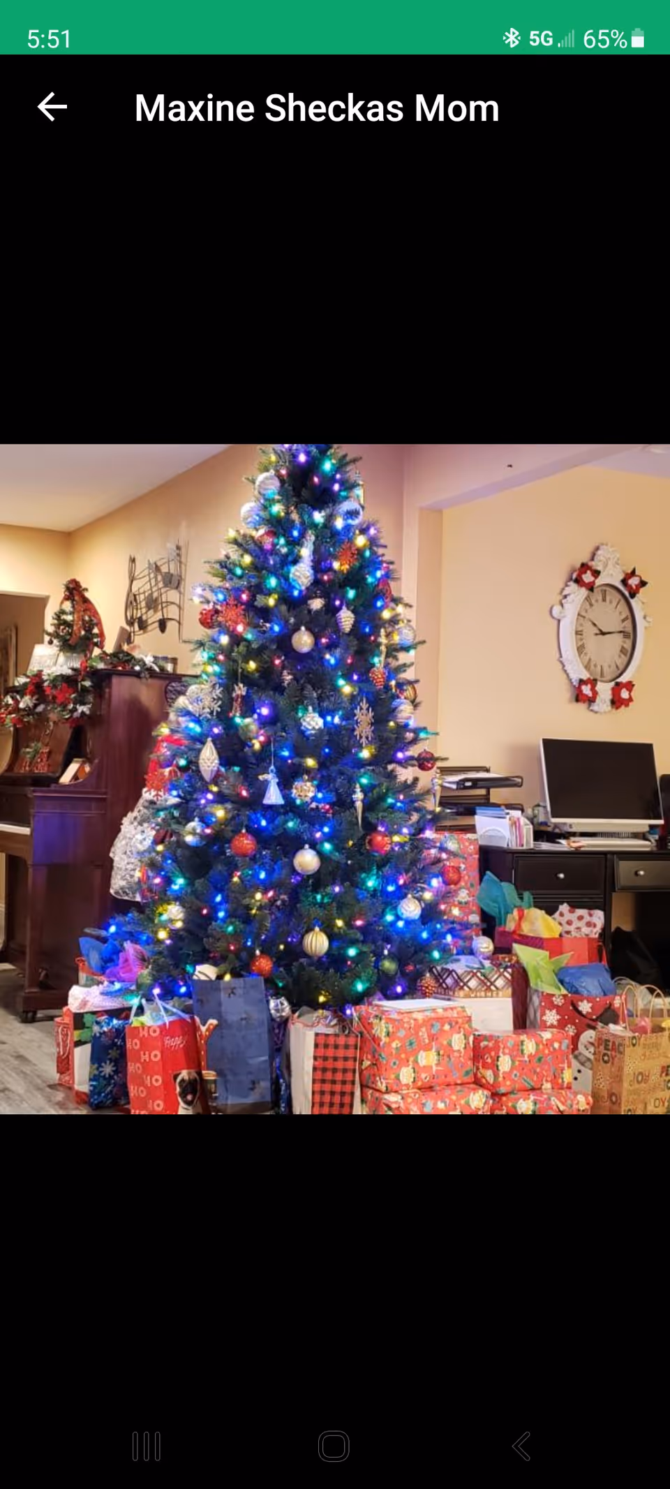 A decorated Christmas tree with colorful lights and ornaments stands in a living room surrounded by numerous wrapped presents. In the background, there is a piano with holiday decorations and a wall clock with a festive wreath around it. A desk with a computer and office supplies is also visible.