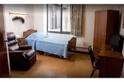 Furnished senior bedroom with a single bed covered by a blue bedspread, a recliner, wooden wardrobe and desk with TV near a window.