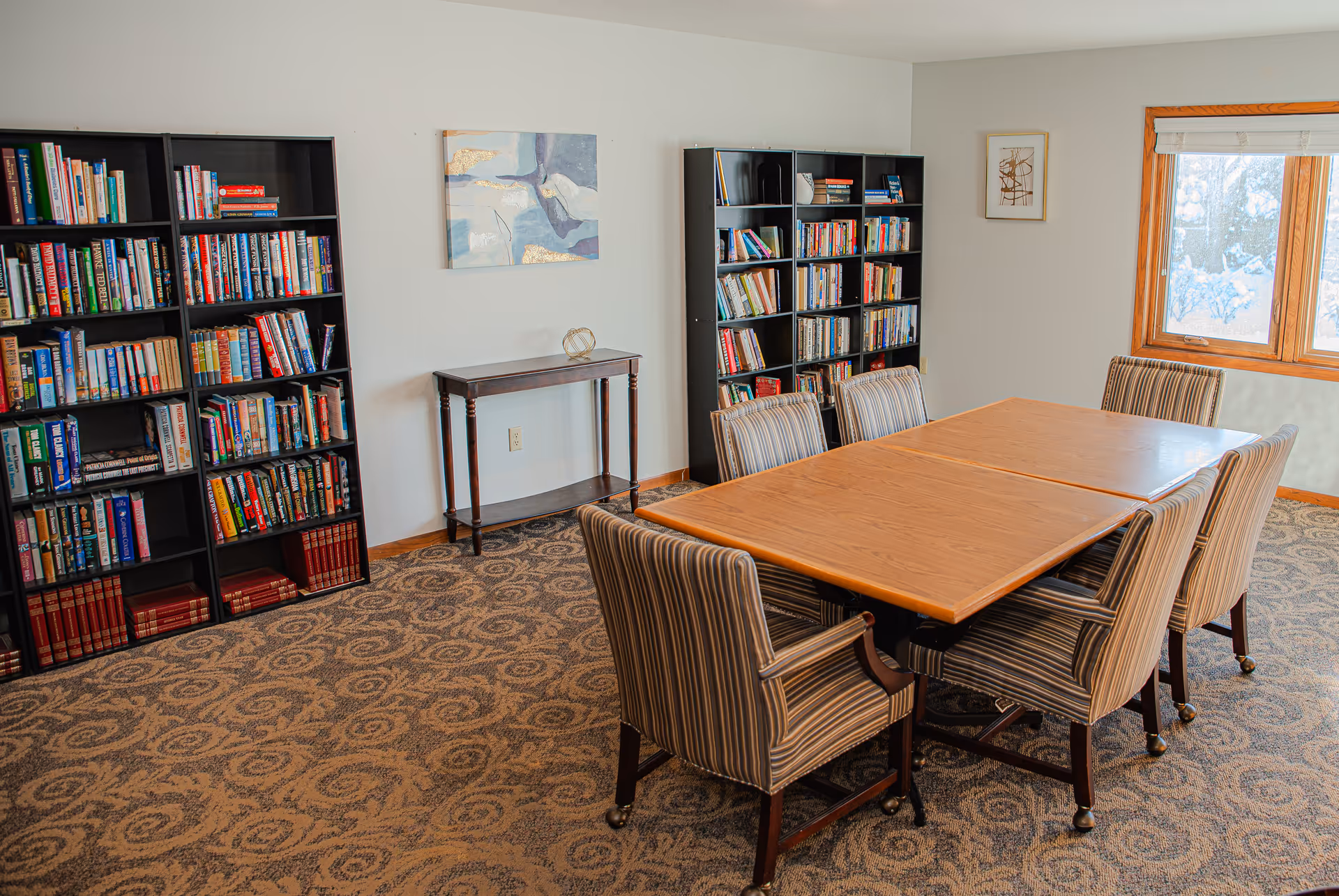 A cozy room with two black bookshelves filled with books, a small wooden table with a decorative item, a large wooden table with six striped upholstered chairs on wheels, patterned carpet, and a window letting in natural light.