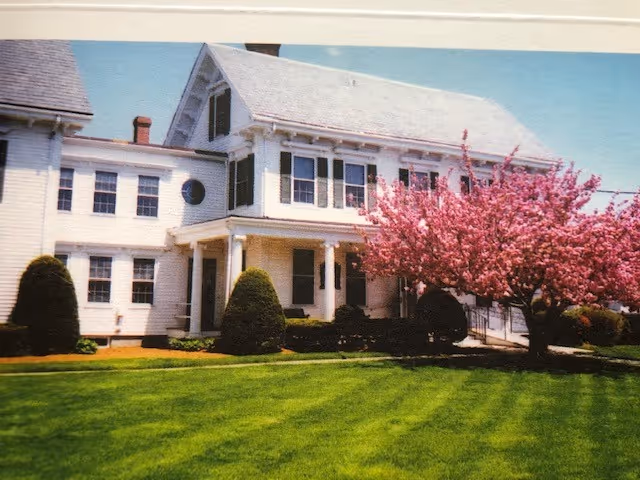 A large white two-story house with black shutters and a covered porch, surrounded by neatly trimmed bushes and a vibrant green lawn. A tree with bright pink blossoms is prominently visible on the right side of the house under a clear blue sky.