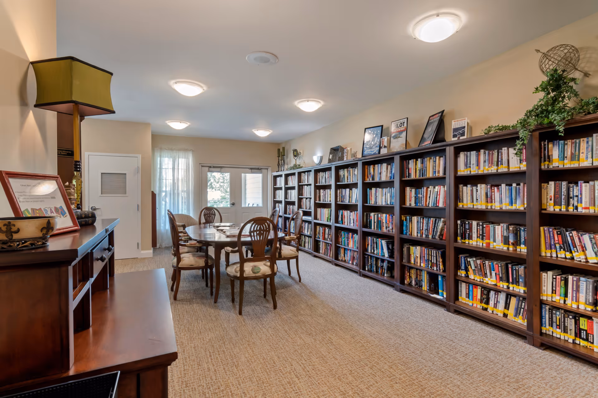 A cozy senior living facility library room with a long wooden table surrounded by six cushioned chairs. The right wall is lined with tall bookshelves filled with books and decorated with framed pictures and a globe with greenery on top. The room has beige walls, carpeted floor, and ceiling lights. A door and window with sheer curtains are visible at the far end.