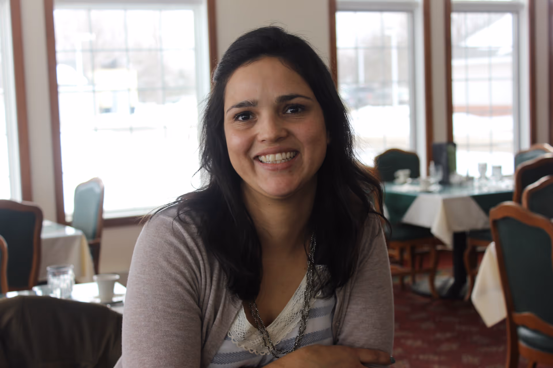 A woman with dark hair smiling and sitting in a dining area with tables and chairs. The room has large windows letting in natural light and is set with tablecloths and dining ware.