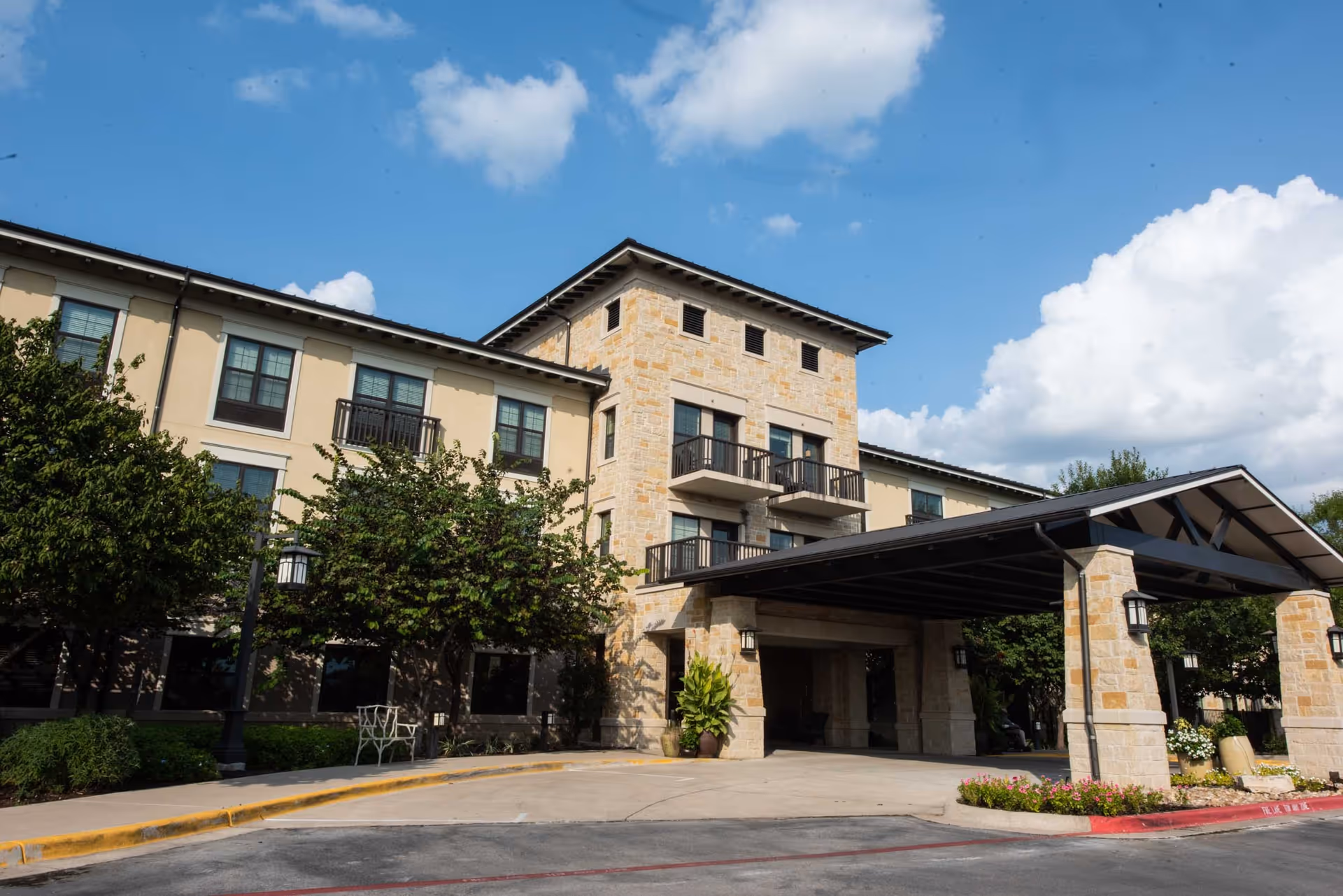 Exterior view of Belmont Village Senior Living West Lake Hills building with a covered entrance, stone and beige facade, trees, and a partly cloudy blue sky.