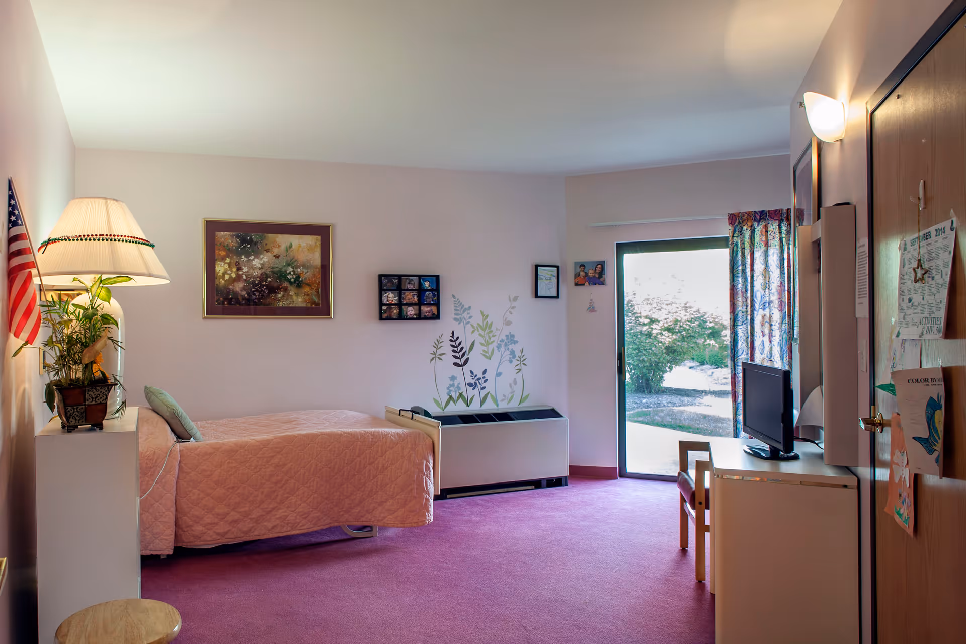 A senior living bedroom with a single bed covered in a pink quilt, a bedside table with a lamp and a plant, an American flag, framed artwork and photos on the wall, a sliding glass door with floral curtains leading outside, a small desk with a TV, and a pink carpeted floor.