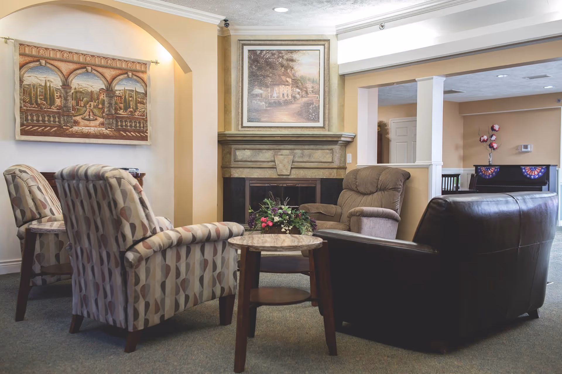 A cozy living room area with a fireplace, featuring two patterned armchairs, a brown recliner, and a dark leather sofa arranged around a small round wooden table with a floral centerpiece. The walls are decorated with a tapestry and a framed painting above the fireplace. In the background, there is a piano with decorative flowers on top.