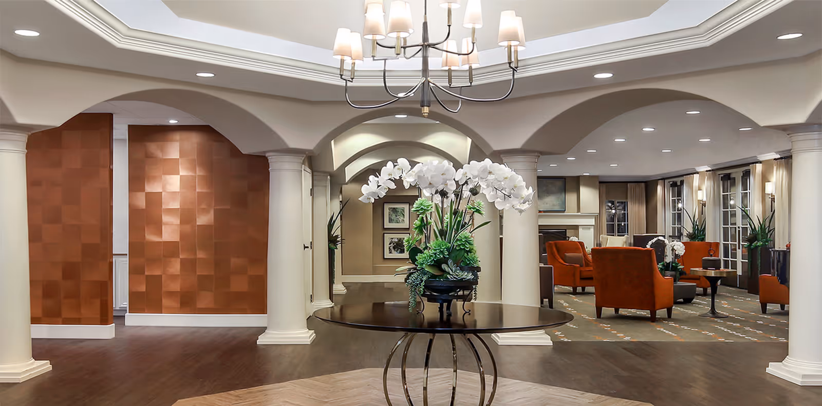 A spacious and elegant senior living facility common area with white columns and arches, a round table with a large floral arrangement in the center, and comfortable orange armchairs arranged around small tables near a fireplace. The room has recessed lighting, large windows with curtains, and a patterned carpet.
