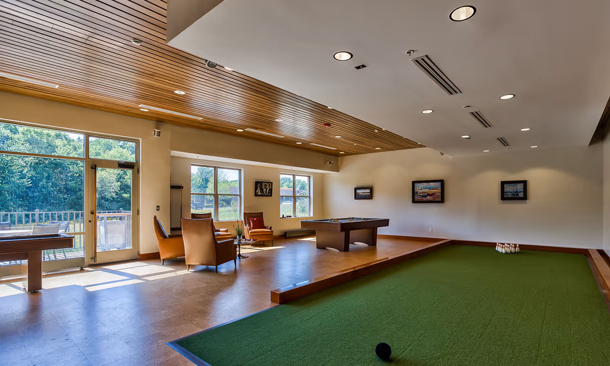 A spacious recreational room with a green indoor bocce ball court in the foreground and a pool table in the background. The room has large windows letting in natural light, several comfortable chairs arranged near the windows, and framed artwork on the walls. The ceiling features wooden paneling and recessed lighting.