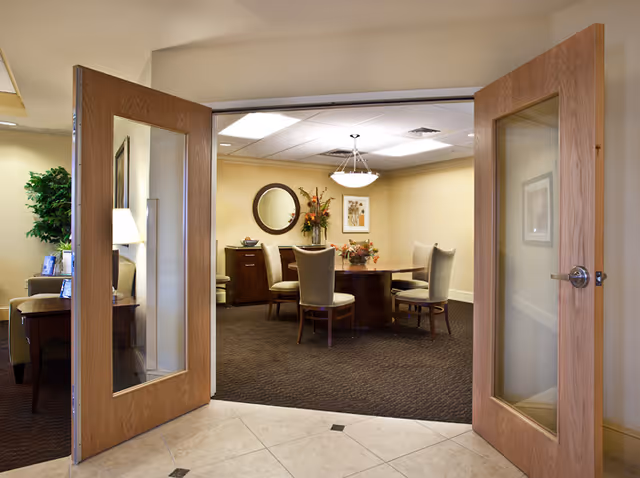 View through open double wooden doors with glass panels into a room with a round table surrounded by six upholstered chairs. The room has beige walls, a dark carpet, a round mirror, framed artwork, and floral arrangements on the table and a sideboard.