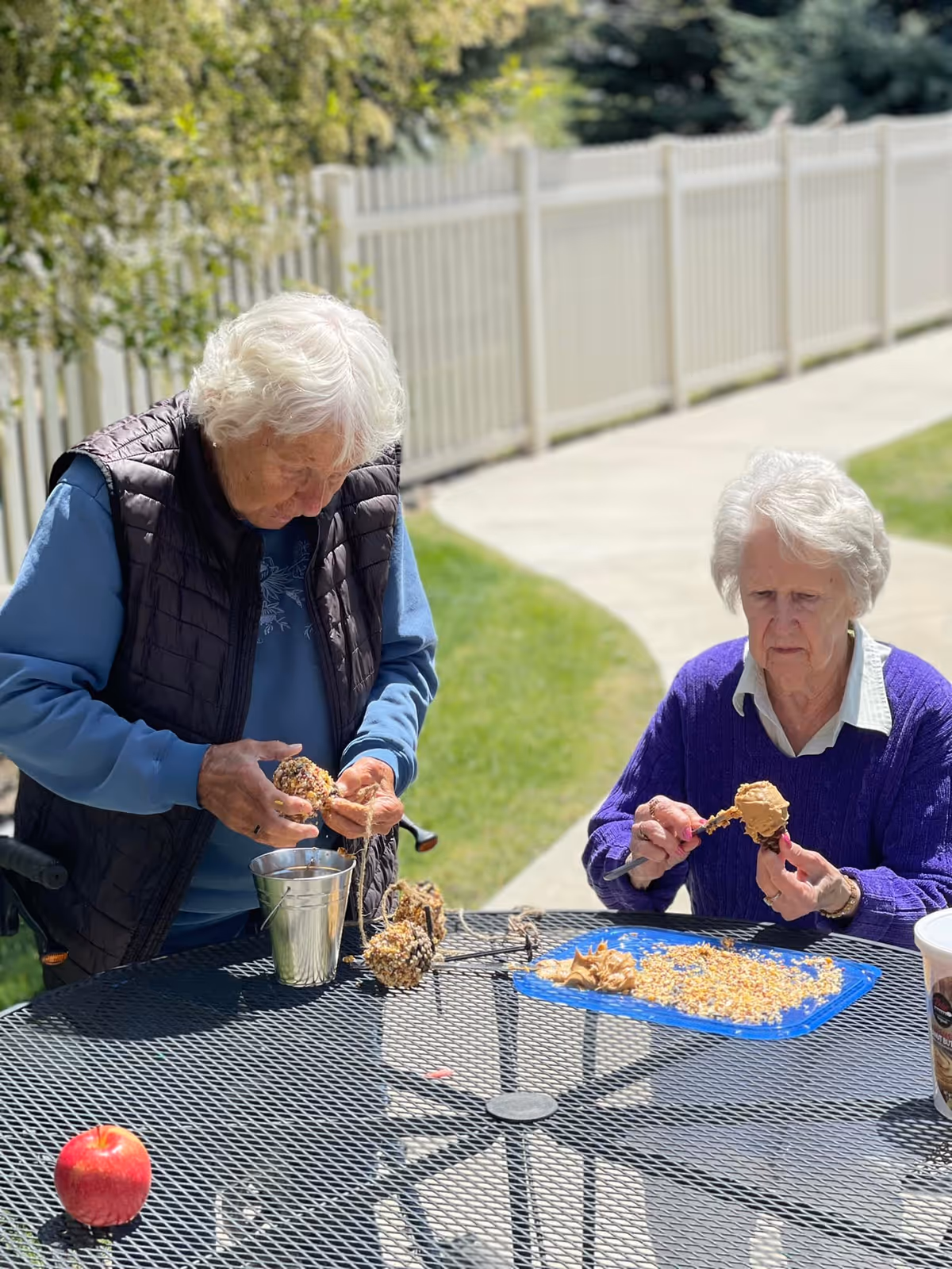 Two elderly women sitting at a black metal outdoor table, making bird feeders by spreading peanut butter on pine cones and rolling them in birdseed. They are outside near a white fence and a concrete pathway, with green grass and trees in the background. One woman is wearing a blue shirt and black vest, the other is wearing a purple sweater over a white collared shirt. An apple and a metal bucket are also on the table.