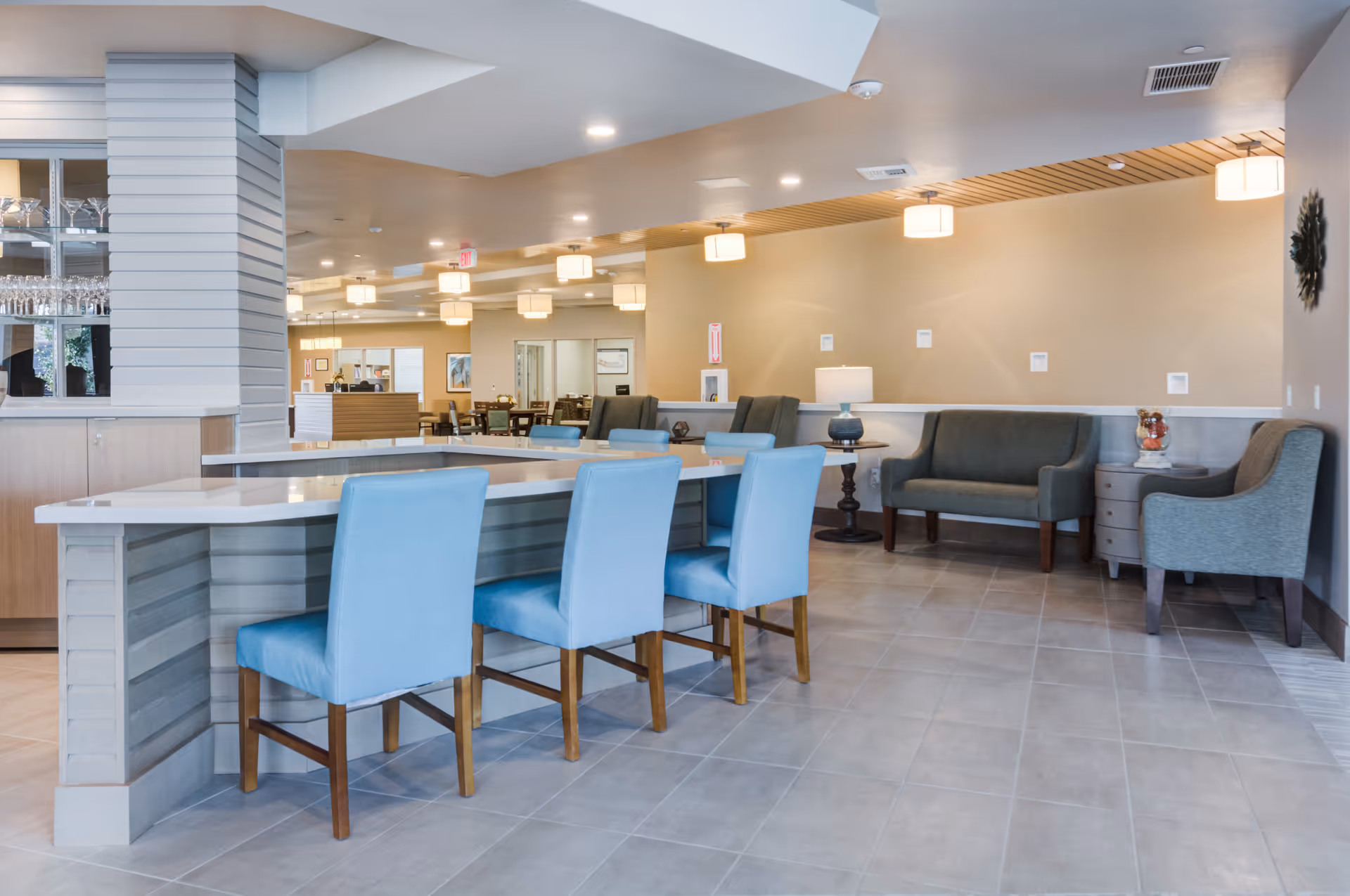A modern senior living facility common area with a long counter and four light blue chairs. In the background, there are additional seating areas with armchairs and a small table with a lamp. The space is well-lit with ceiling lights and has a clean, neutral color palette.