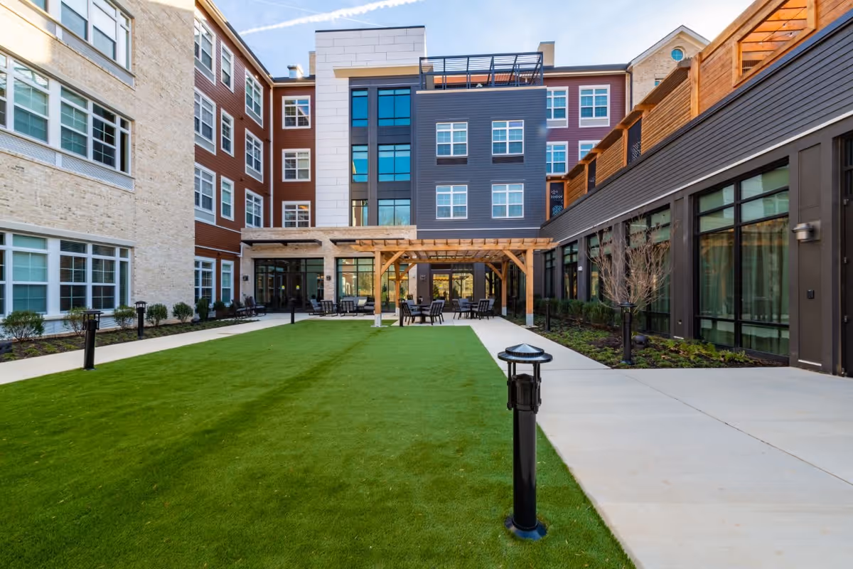 Outdoor courtyard area of Tribute at Black Hill facility featuring a well-maintained green lawn, concrete walkways, modern black outdoor lights, and a wooden pergola with seating underneath. The multi-story building surrounds the courtyard with large windows and a mix of brick, siding, and panel exteriors under a clear blue sky.
