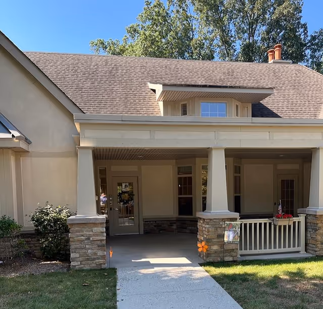 Front entrance of a senior living facility with a covered porch supported by columns with stone bases, a concrete walkway leading to the door, and some greenery on the sides. The building has a beige exterior and a shingled roof with a small dormer window.