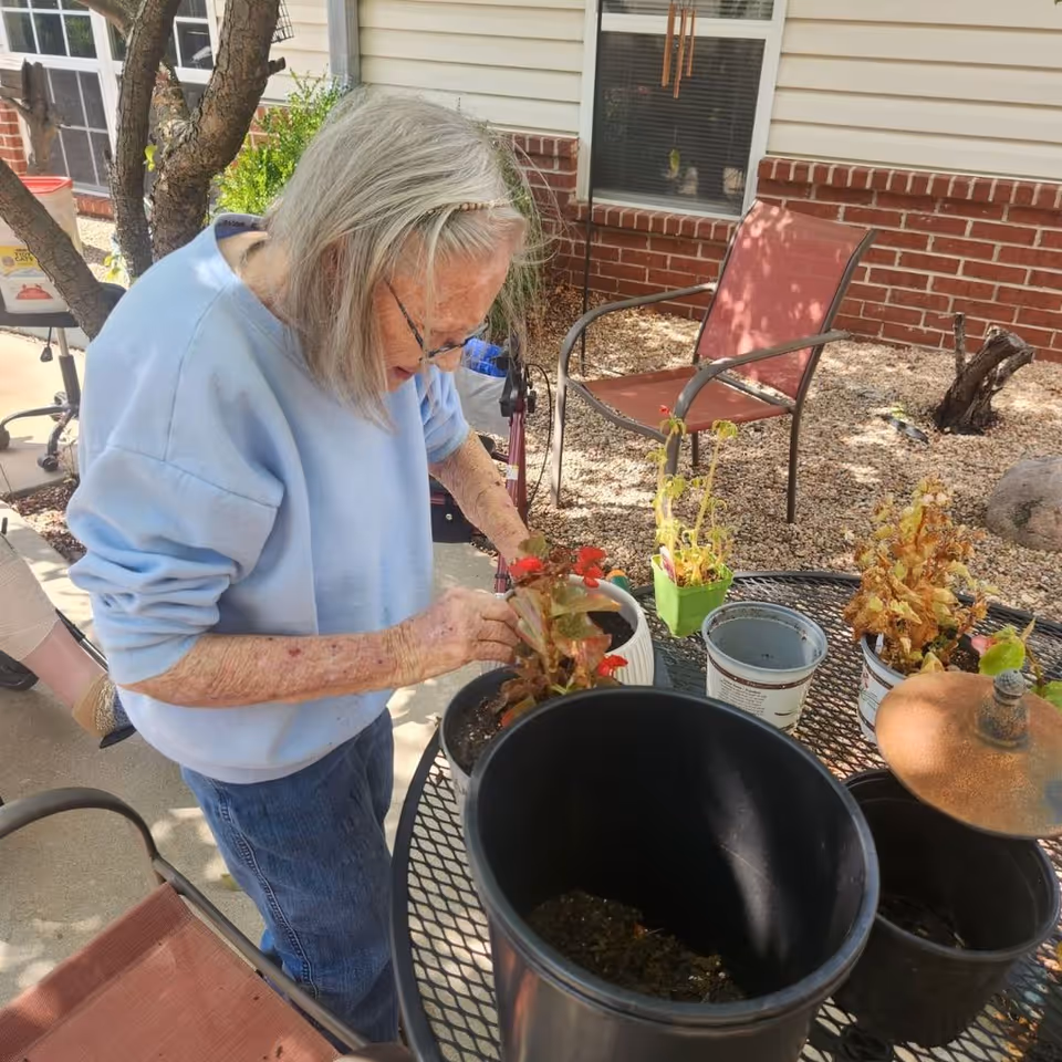An elderly woman with gray hair and glasses is tending to potted plants on a round metal table outdoors. She is wearing a light blue sweatshirt and jeans. The background shows a brick building with windows, a red patio chair, and some gravel landscaping with small trees and plants.